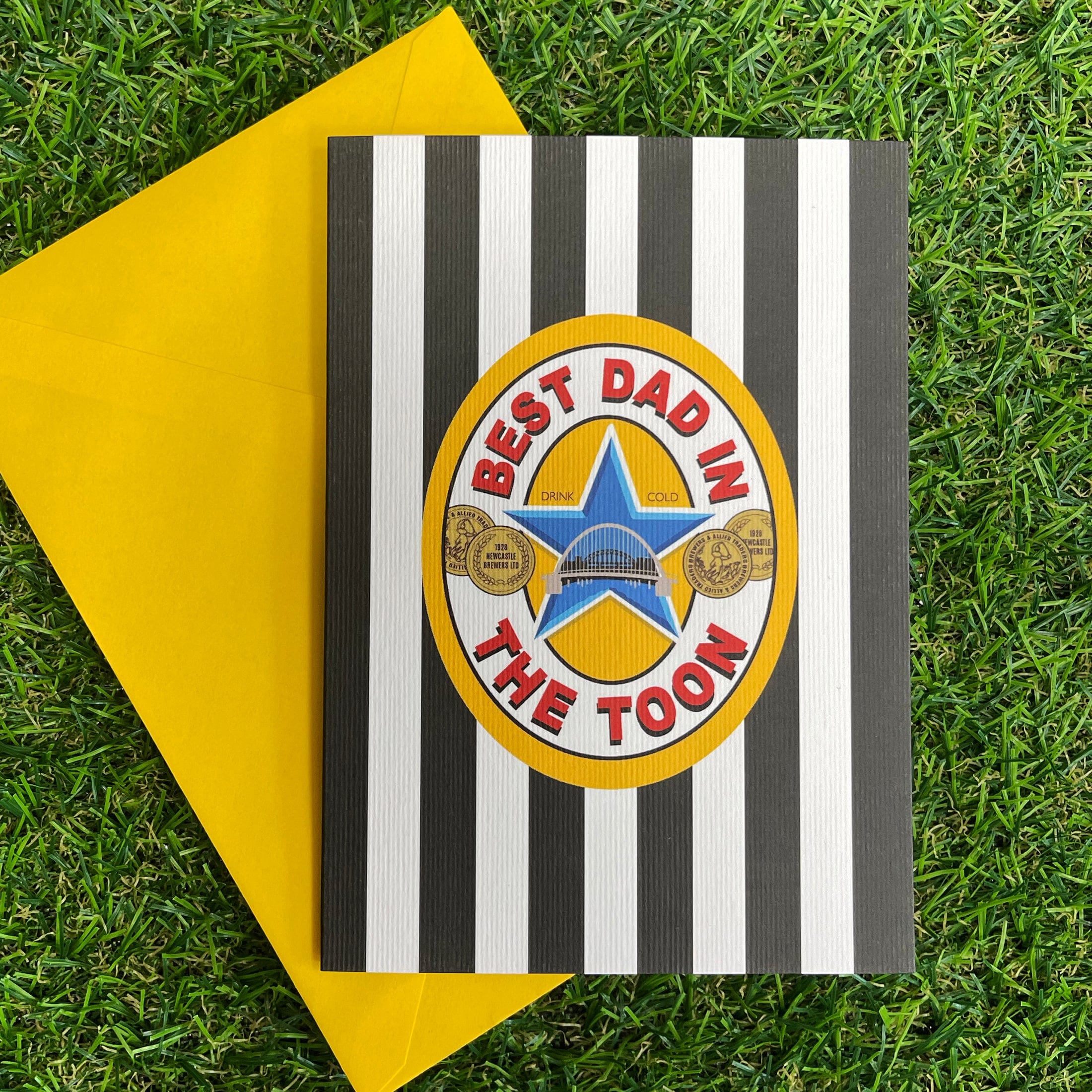 Black-and-white striped greeting card with “Best Dad in The Toon” badge design, shown with a yellow envelope on green grass background.