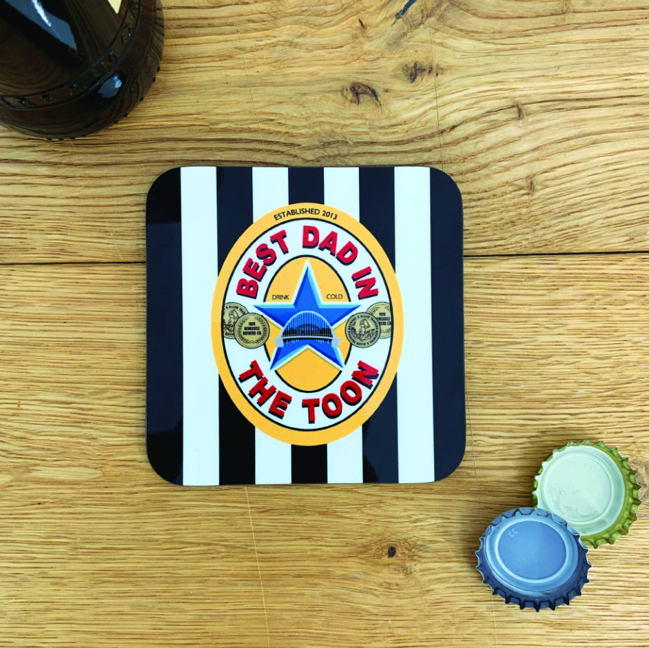 Black-and-white striped coaster reading “Best Dad in The Toon” with star and bridge design on wooden table.