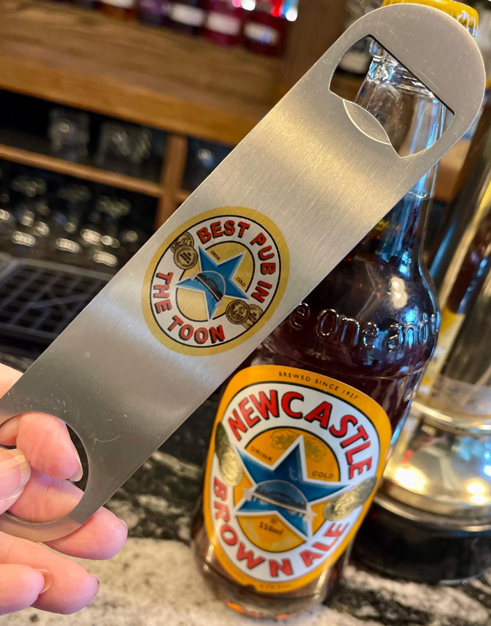 Close-up of a stainless steel bottle opener with “Best Pub in The Toon” design, held beside a bottle of Newcastle Brown Ale at a bar.