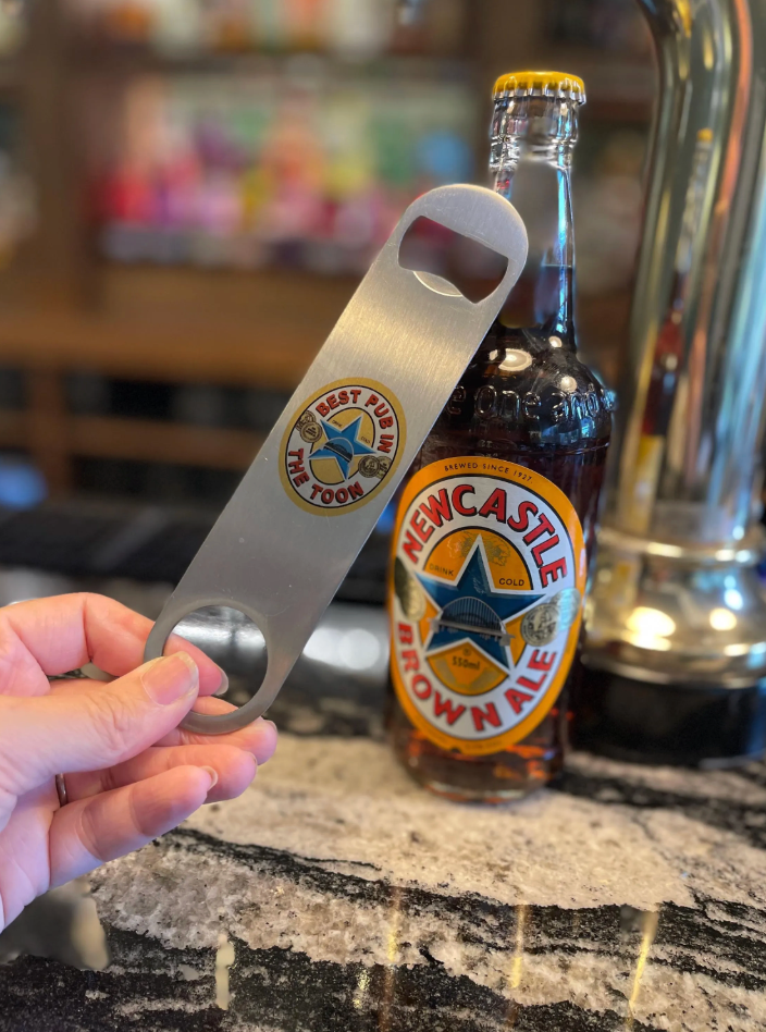 Hand holding a Newcastle Brown Ale bottle opener with a bottle of Newcastle Brown Ale in the background.