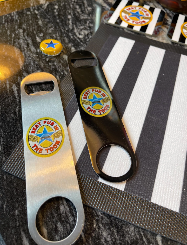 Silver and black “Best Pub in The Toon” bar blade openers displayed on a black-and-white striped surface beside bottle caps — perfect gift for football fans and home bars.