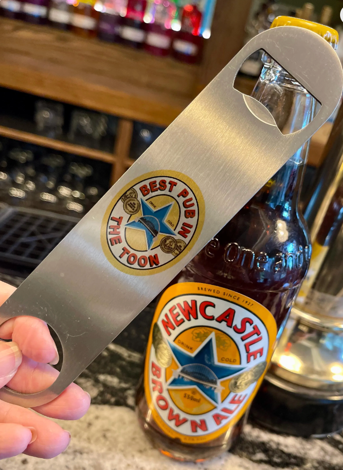 Close-up of a stainless steel bottle opener with “Best Pub in The Toon” design, held beside a bottle of Newcastle Brown Ale at a bar.